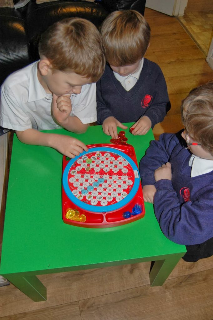 Three young children in school uniforms engage in a friendly competition, playing a fishing game with magnetic rods at a green table. They focus intently on catching small fish pieces from a red and blue game board.