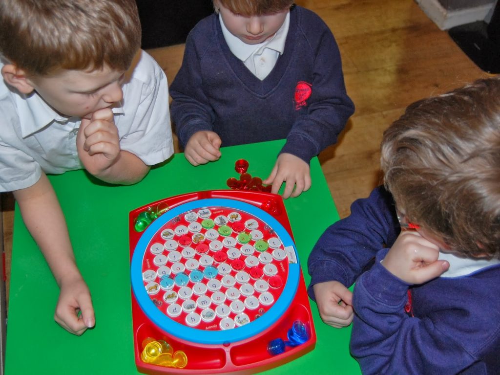 Three young children in school uniforms sit around a green table, playing a colourful board game similar to Wordsearch Junior, visibly focused and engaged as if in friendly competition.