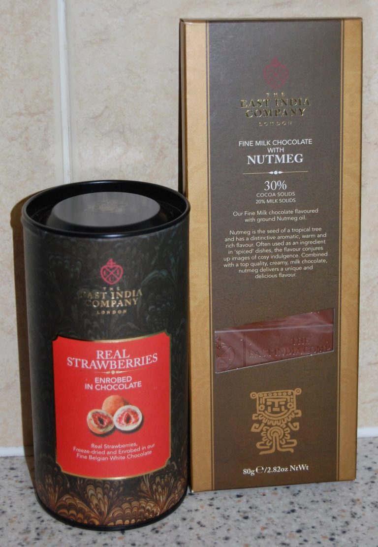 A cylindrical container of East India Company real strawberries coated in chocolate from The East India Company stands next to a box of fine milk chocolate with nutmeg. Both packages are upright on a light-coloured surface.