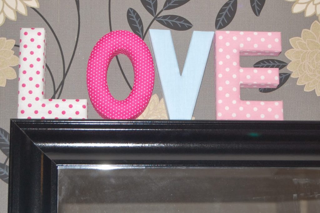 Soft decorative letters from Wilko spelling "LOVE" in different pastel shades and patterns are displayed on top of a black-framed mirror, with floral patterned wallpaper in the background—perfect for unique Valentine’s Day gifts.