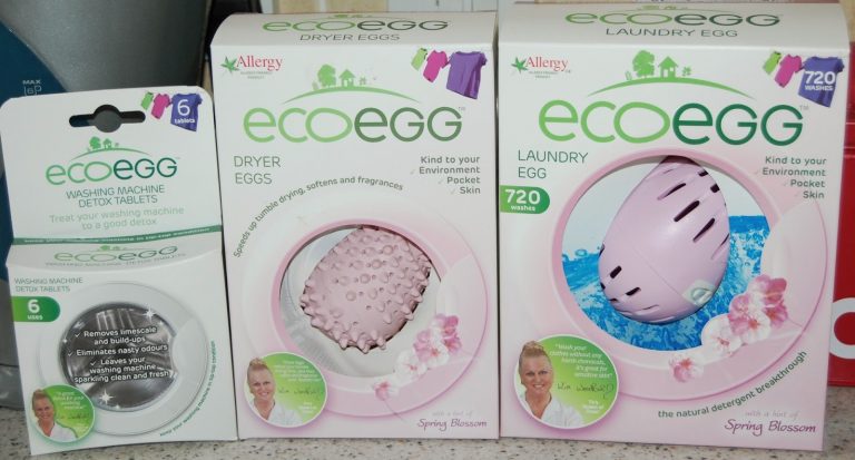 Three Ecoegg products on a counter: the Laundry Egg, Washing Machine Detox Tablets, and Dryer Eggs in Spring Blossom scent. Packaging is white and purple, featuring eco-friendly claims, a woman's photo, and hints of competition.