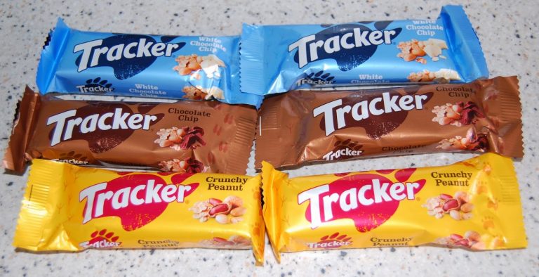 Six Tracker snack bars are displayed on a worktop: two blue (White Chocolate Chip), two brown (Chocolate Chip), and two yellow (Crunchy Peanut) wrappers, arranged in two rows of three—perfect for a quick Tracker review.