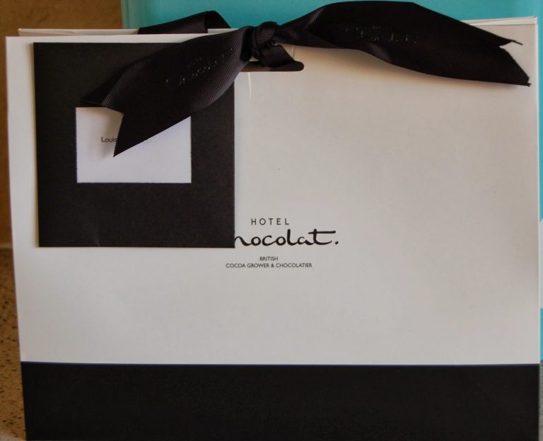 A white Hotel Chocolat gift bag with a black ribbon bow on top and a black envelope attached, featuring the Hotel Chocolat logo in the centre—a perfect treat for mum.