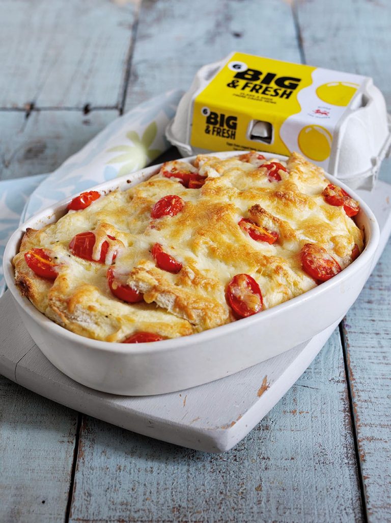 A baked egg casserole made with fresh eggs is topped with melted cheese and sliced cherry tomatoes in a white dish. Behind it, a "Big & Fresh" carton rests on a rustic wooden surface—perfect inspiration for your next eggs recipe.