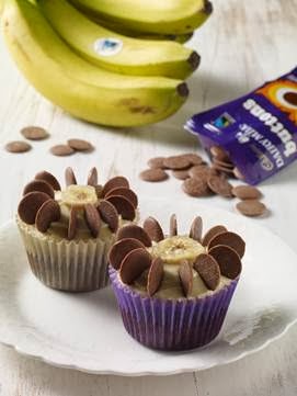 Two banana flower cupcakes topped with banana slices and Fairtrade chocolate buttons sit on a white plate. In the background, a bunch of bananas and a packet of chocolate buttons rest on a light wooden surface.
