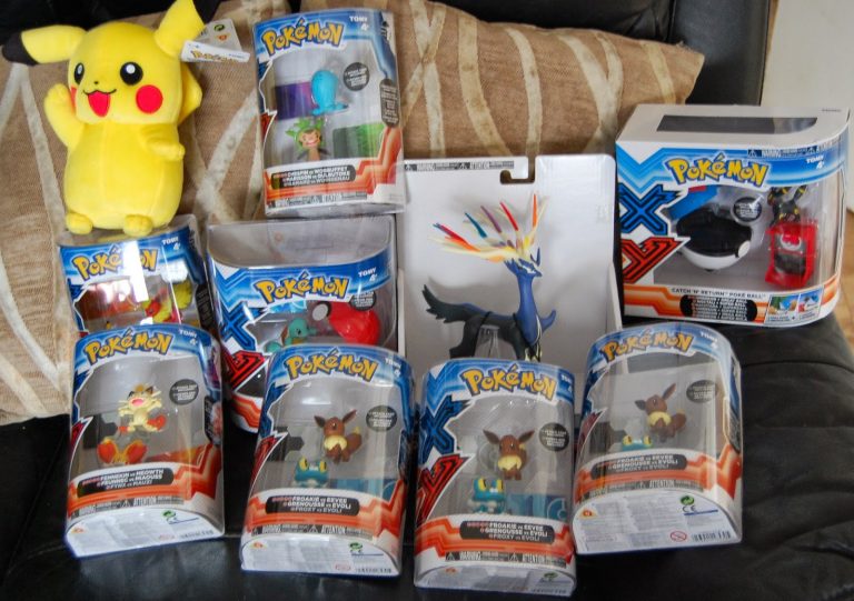 Several Pokémon action figures in packaging, a boxed set from the Pokémon X and Y range of toys, and a yellow Pikachu soft toy are displayed on a black sofa with beige striped cushions in the background.