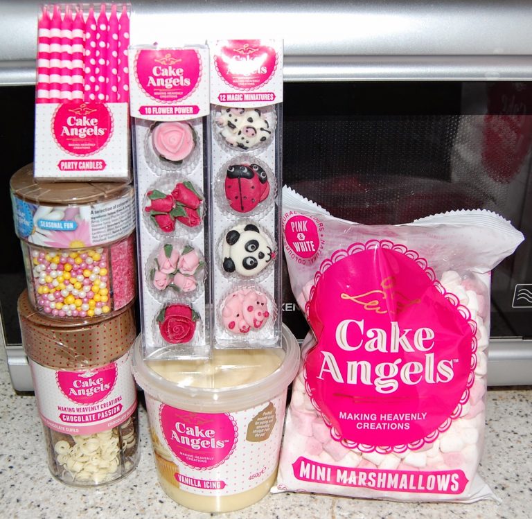 A selection of Cake Angels baking products, including party candles, colourful sprinkles, flower and ladybird cake decorations, vanilla icing, and a bag of pink and white mini marshmallows, displayed in front of a microwave oven.