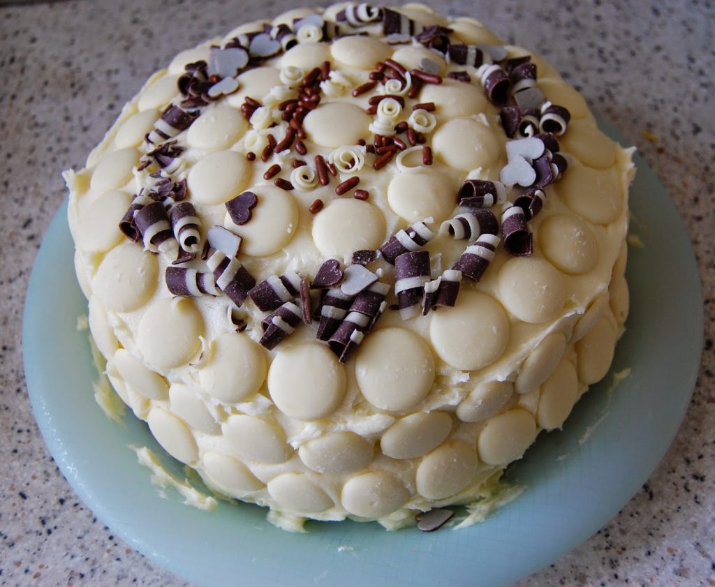 A round cake covered in white chocolate buttons, adorned with Cake Angels cake decorations like chocolate curls, hundreds and thousands, and heart-shaped pieces on top, sits on a light blue plate.