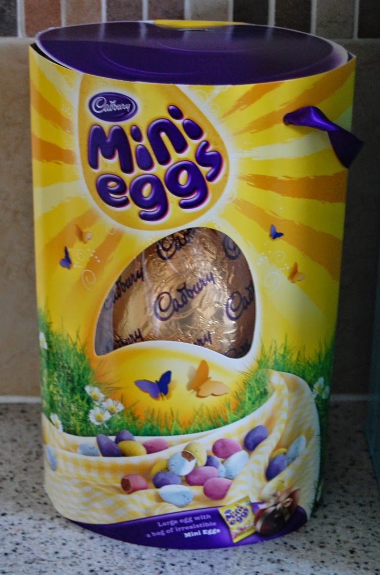 A large, yellow cylindrical box of Cadbury Mini Eggs with a purple lid sits on a worktop. The Easter-themed design features colourful Easter Eggs, butterflies, and a visible chocolate egg inside—perfect for your Ocado basket.
