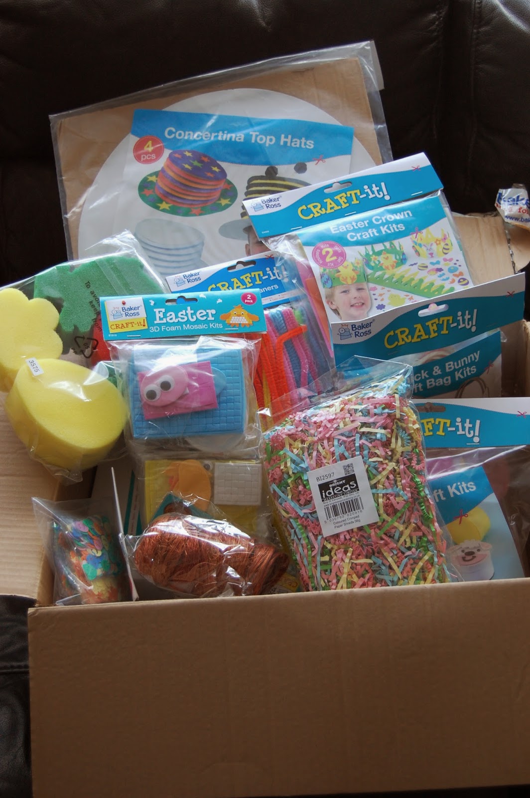 A Baker Ross cardboard box filled with colourful, crafty supplies—including Easter-themed kits, foam shapes, paper hats, rainbow shredded paper, pipe cleaners, and packaged wool.