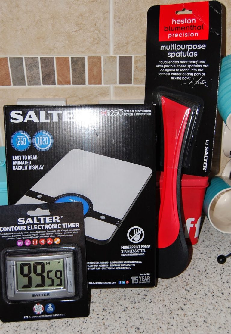 A boxed Salter digital kitchen scale, a red Heston Blumenthal multipurpose spatula in packaging, and a Salter electronic timer displaying 99:59 on a kitchen worktop—perfect for your next Salter Challenge or making an Easter dessert.