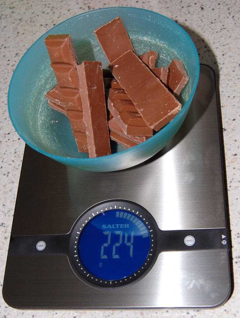 A blue bowl filled with pieces of milk chocolate, perfect for the Chocolate Easter Nest Challenge, sits on a digital kitchen scale reading 224 grams. The scale rests on a speckled worktop.