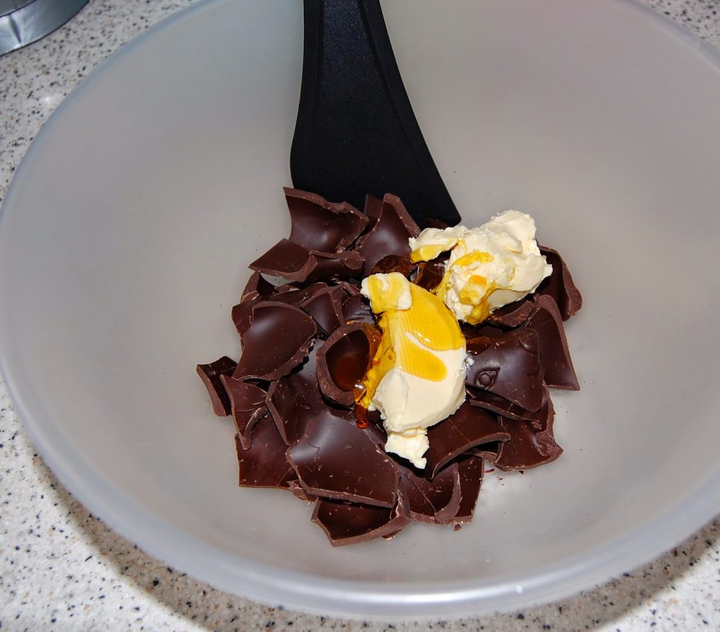 A mixing bowl containing broken pieces of dark chocolate, a knob of butter, and a small amount of golden syrup, with a black spatula resting in the bowl—perfect for your next Chocolate Easter Nest creation.