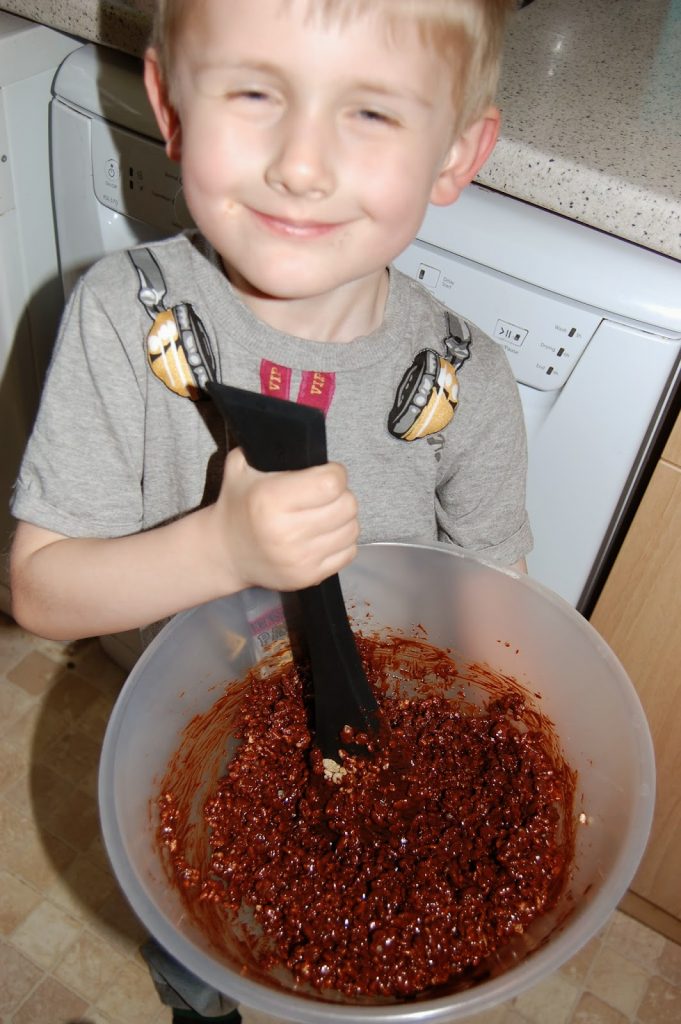 Smiling child holding a mixing bowl filled with chocolate and Rice Krispie mixture, stirring with a black spatula in a kitchen—creating a delightful Chocolate Easter Nest for an Easter dessert Salter Challenge.