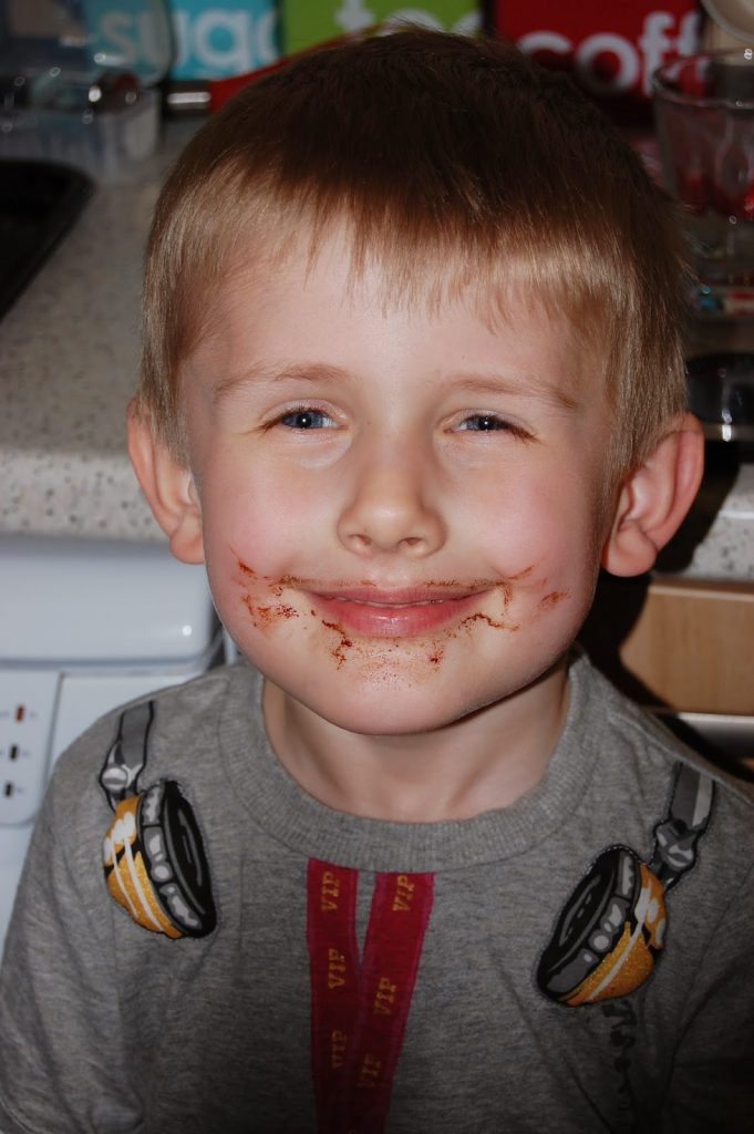 A young boy with fair hair and blue eyes smiles with Chocolate Easter Nest smeared around his mouth. He is wearing a grey shirt with a headphone graphic and is standing in a kitchen, looking proud of his Easter Challenge.