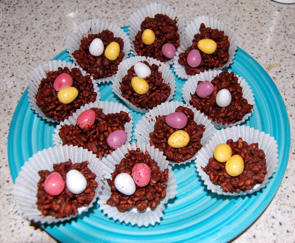 A blue Salter plate holds chocolate rice crispy nests in white paper cases, each topped with two or three colourful candy eggs—a sweet entry for the Chocolate Easter Nest Challenge on a speckled worktop.