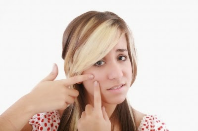 A young woman with long, blonde-highlighted hair is touching her cheek and pointing at spots on her face, looking concerned. She is wearing a white top with red polka dots.