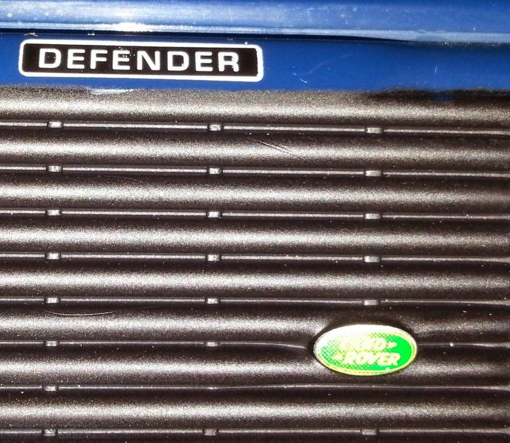 Close-up of a vehicle grille with a "DEFENDER" badge at the top and a green Land Rover logo near the bottom, set against a dark, ribbed surface—perfect inspiration for collectors of big farm toys and rugged models.