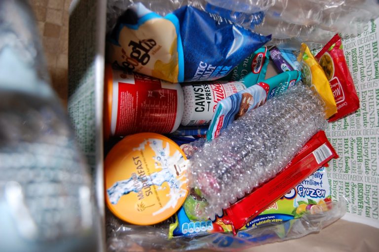 A Degustabox filled with assorted snacks and food items, including crisps, a tin, chocolate bars, a round orange container, and a bottle wrapped in bubble wrap—all packed with plastic protective lining. Perfect for an April review!.