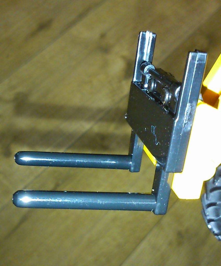 Close-up of the front fork attachment on a Big Farm Toys forklift, showing two black plastic prongs extended outward, with a wooden floor in the background.