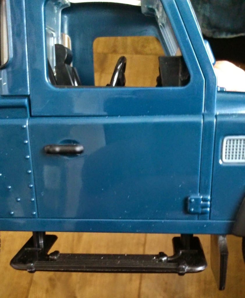 Close-up of a blue Big Farm Toys lorry's side with an open door, visible steering wheel, and black footstep. The shiny lorry sits on a wooden floor.