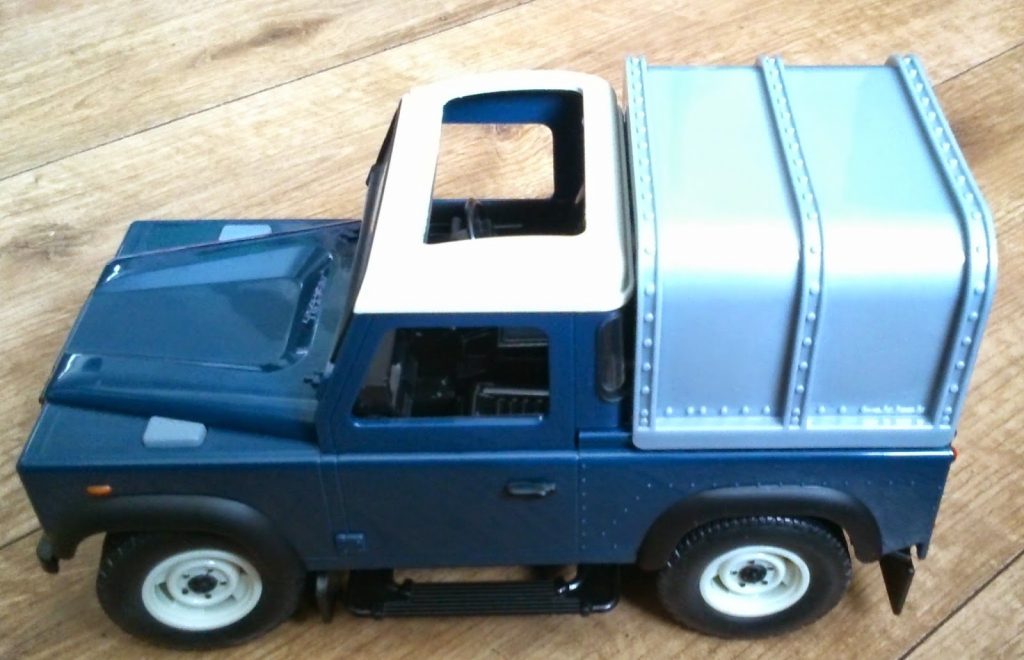 A toy model of a blue and white pick-up truck with a silver, boxy cargo bed is displayed on a wooden surface. Perfect for fans of Big farm toys, it features a sunroof, detailed wheels, and wing mirrors.