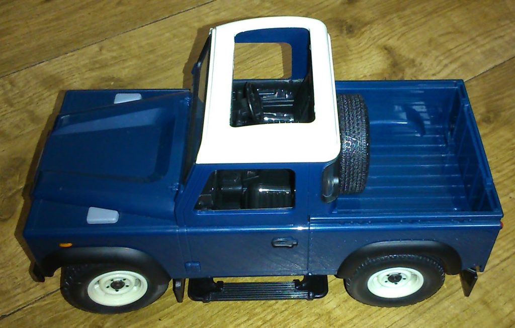 A blue toy pickup truck from Big Farm Toys with a white roof and black tyres sits on a wooden floor. The truck features an open bed and a spare tyre mounted inside the bed.