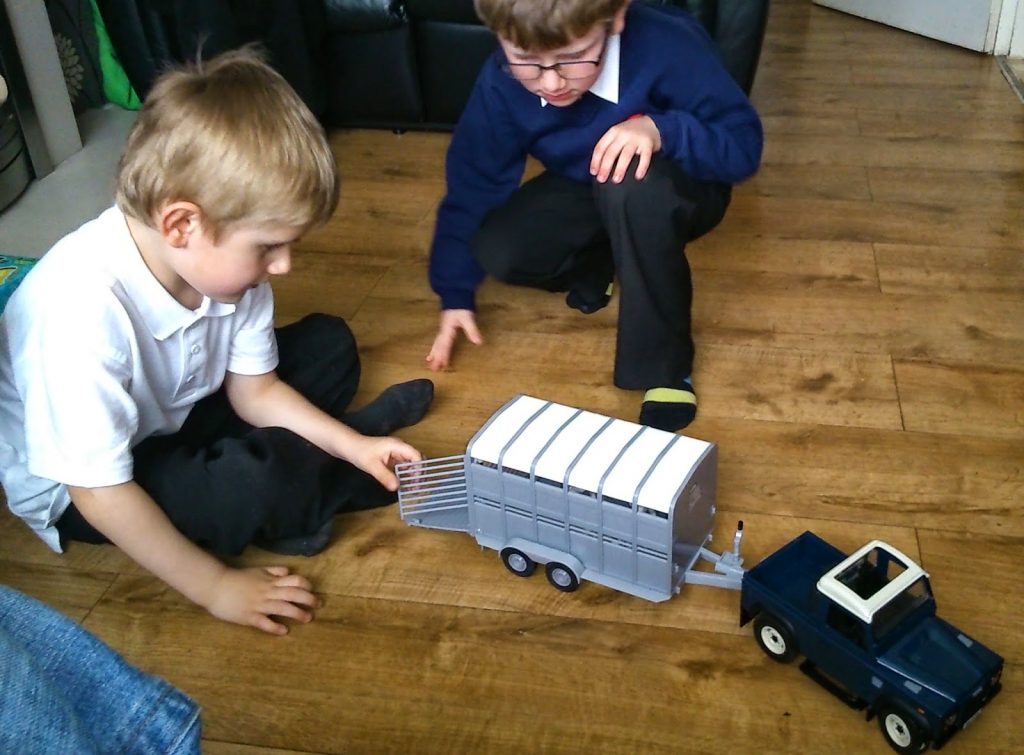 Two young boys play on a wooden floor with a toy blue pick-up truck and silver trailer, just like Big Farm Toys. One boy opens the trailer gate whilst the other kneels nearby, both focused on their imaginative play.