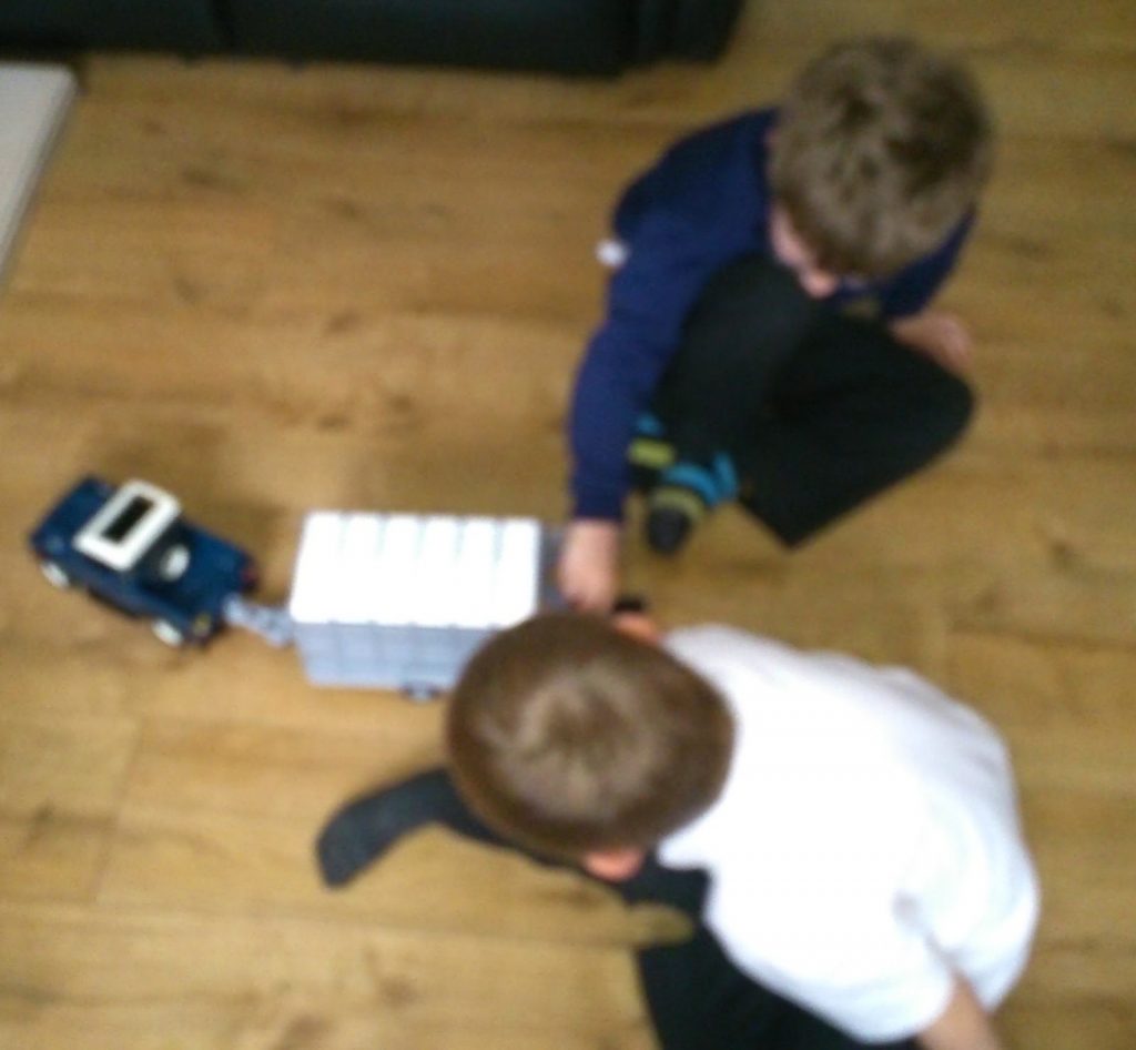 Two children sit on a wooden floor, playing with a toy lorry from their collection of large farm toys. The focus is on their interaction with the toy, and their faces are not clearly visible.