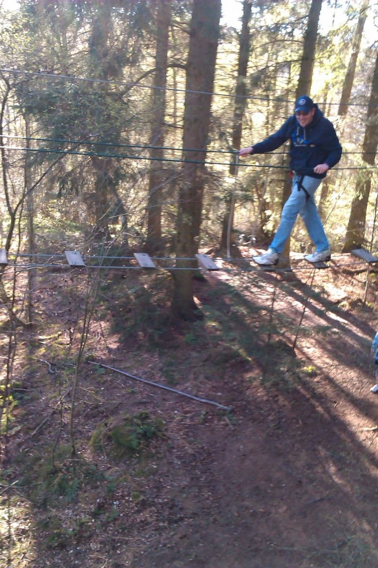 A person wearing a safety harness crosses a rope bridge with wooden planks in a Go Ape adventure park, surrounded by tall trees and sunlight filtering through the leaves.