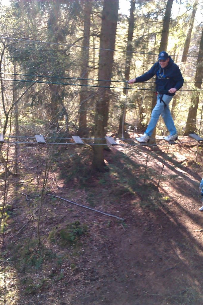 A person wearing a harness and helmet carefully crosses a suspended rope bridge with wooden planks at a Go Ape Treetop Adventure park, whilst sunlight filters through the tall trees.