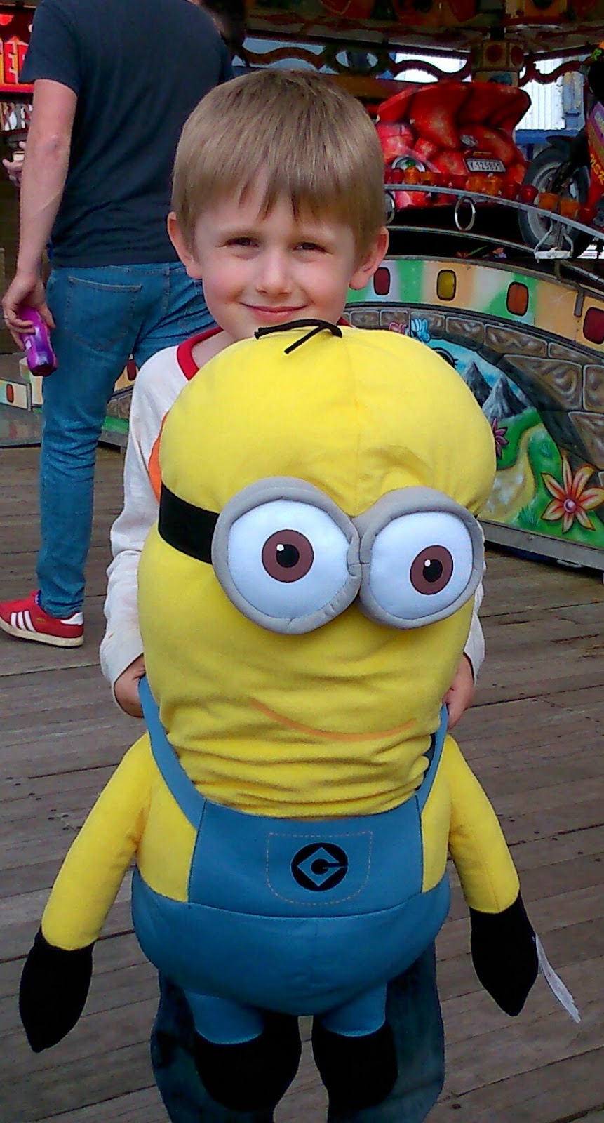 A smiling young boy stands on a wooden boardwalk holding a large Minion soft toy in front of him at an amusement park, with brightly coloured rides, another person, and signage for Madame Tussauds visible in the background.