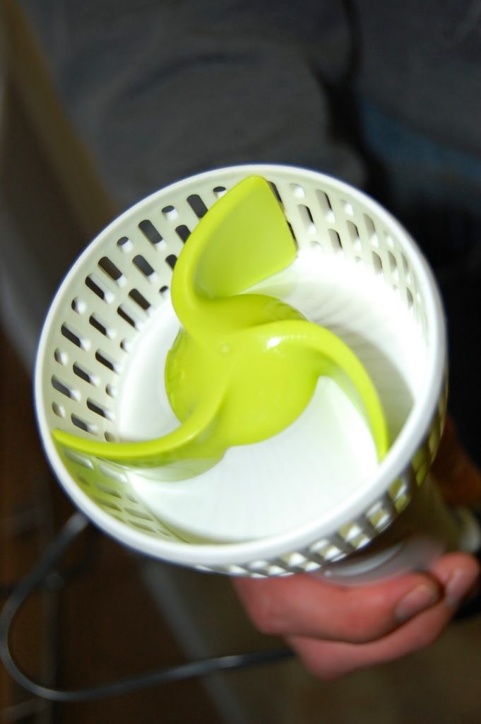A close-up of a hand holding a white plastic container with a green three-blade spinning attachment inside, likely part of a Masha appliance designed for creating the perfect mash in your kitchen.