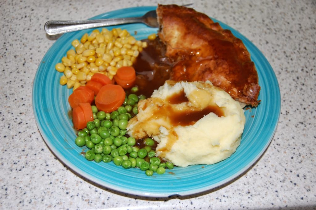 A blue plate with Perfect Mash topped with brown gravy, garden peas, sliced carrots, sweetcorn, and a piece of meat pie, with a fork at the side.
