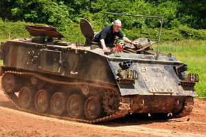 Two people ride in an old, open-topped military tracked vehicle on a dirt road, surrounded by greenery. The vehicle kicks up dust as it moves—an adventurous outing that could inspire unique Father’s Day gift ideas.