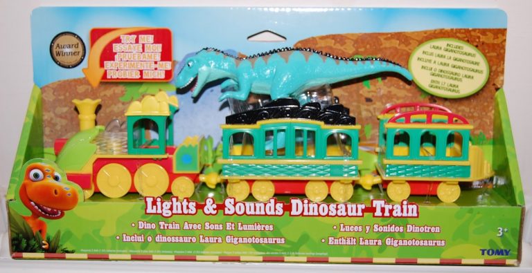 A colourful Dinosaur Train toy set packaged in a box, featuring a blue dinosaur figure on the train cars. The box reads “Lights & Sounds Dinosaur Train” and displays text in multiple languages, with a cartoon dinosaur and TOMY logo.