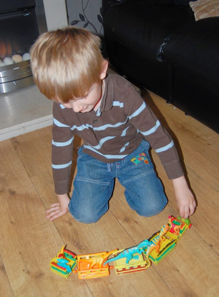 A young child with light brown hair, wearing a brown striped shirt and blue jeans, kneels on a wooden floor whilst playing with a colourful Dinosaur Train set.