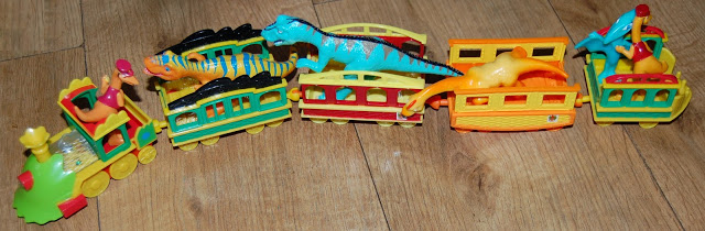 A colourful Dinosaur Train with four carriages, each carrying plastic dinosaur figures, sits on a wooden floor. The train is yellow, green, and red, and the dinosaurs come in various colours and sizes.