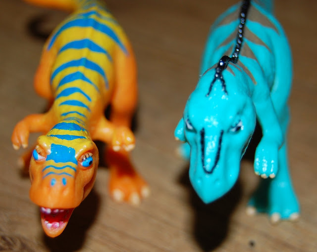 Two toy dinosaurs, inspired by Dinosaur Train—one orange with blue stripes and the other teal with black stripes—stand on a wooden surface, both facing forwards.