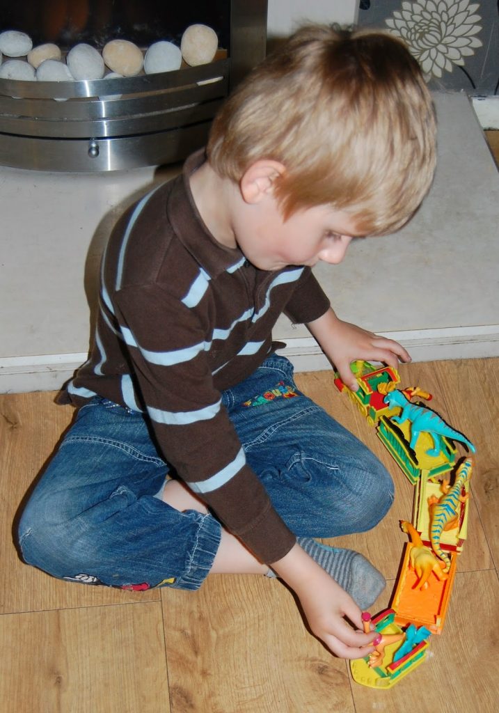 A young boy with blond hair, wearing a brown and blue striped shirt and jeans, sits cross-legged on a wooden floor playing with a colourful Dinosaur Train toy set.