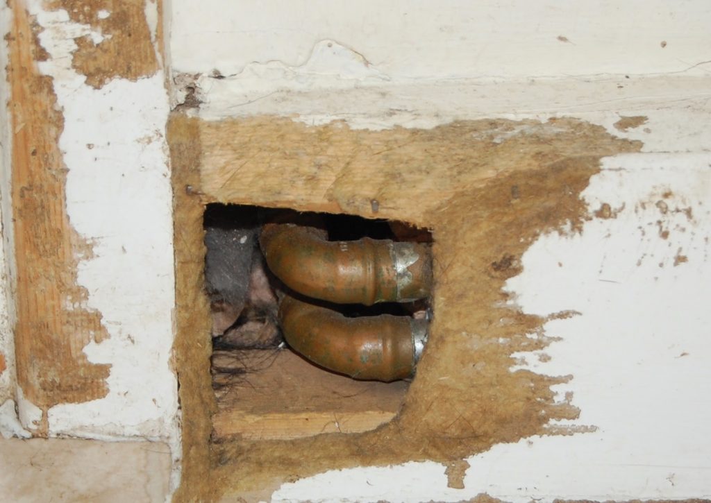 A close-up of two metal pipes running through a rough hole cut into a white painted wooden wall, with chipped paint and exposed wood edges visible—almost as if revealing how naughty your pet is in their wildest antics.
