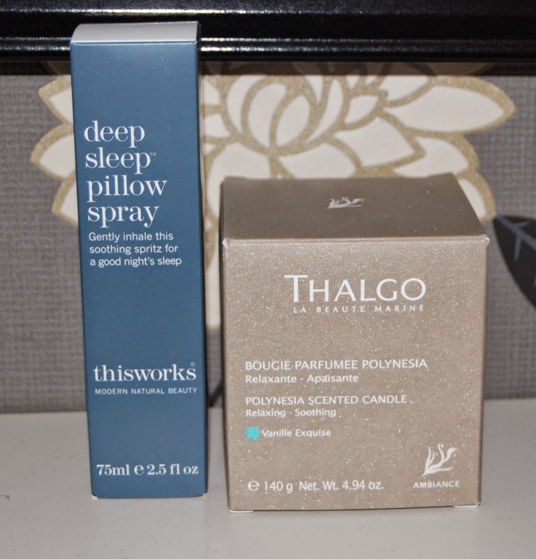 A box of "deep sleep pillow spray" by thisworks, designed to help you enjoy a good night's sleep, sits beside a gold Thalgo Polynesia scented candle, both arranged on a flat surface against a patterned background.