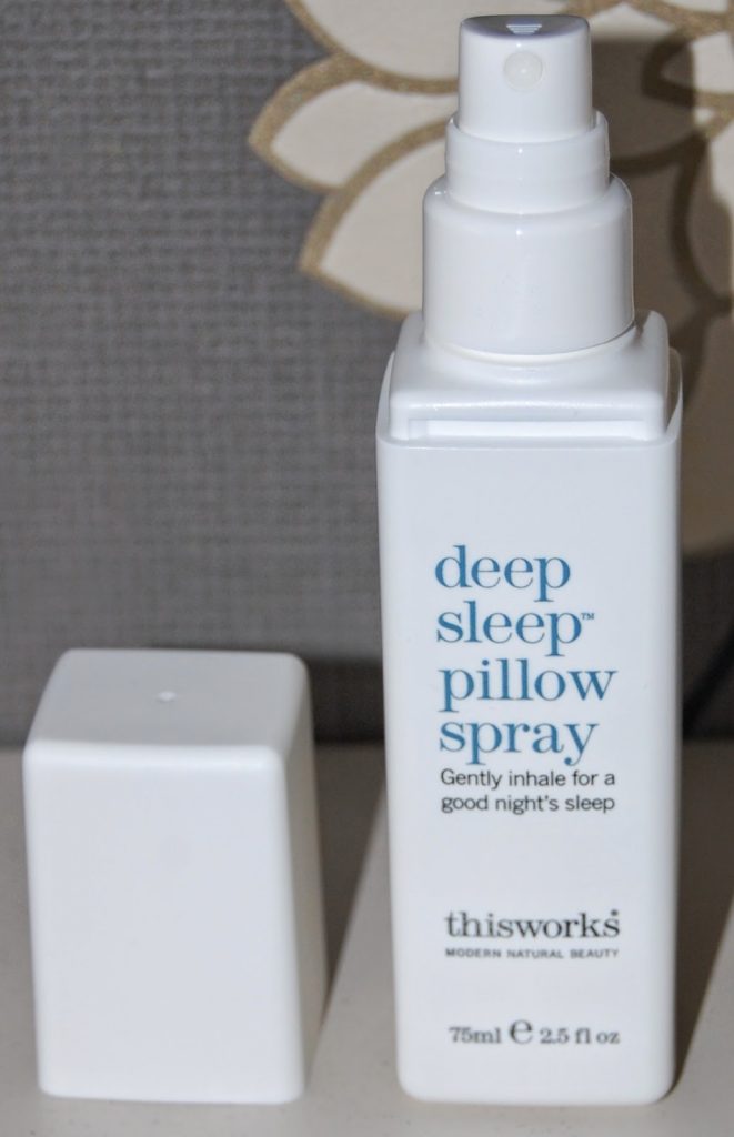 A white bottle of "deep sleep pillow spray" by thisworks with its cap removed, standing on a surface. The label suggests gently inhaling for restful sleep and a good night’s sleep.
