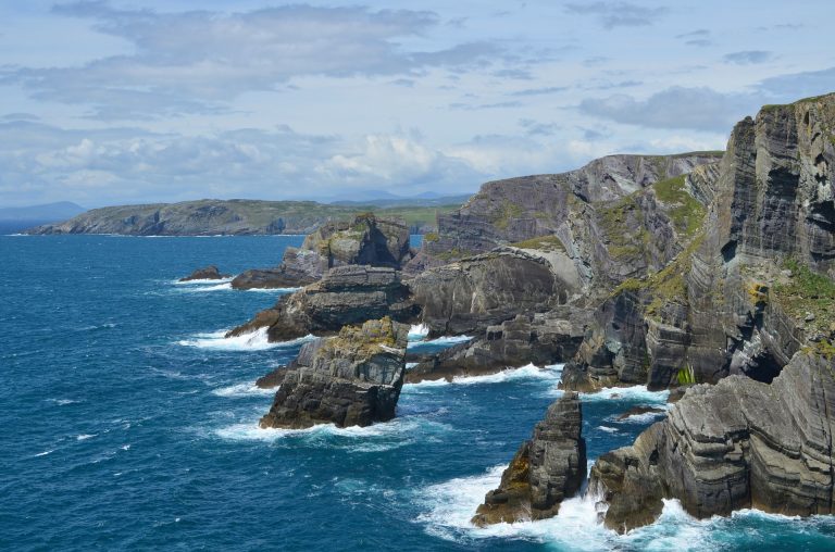 Dramatic rocky cliffs rise above the blue sea with waves crashing at their base under a partly cloudy sky, forming a rugged Cork coastline in Ireland with distant hills in the background.
