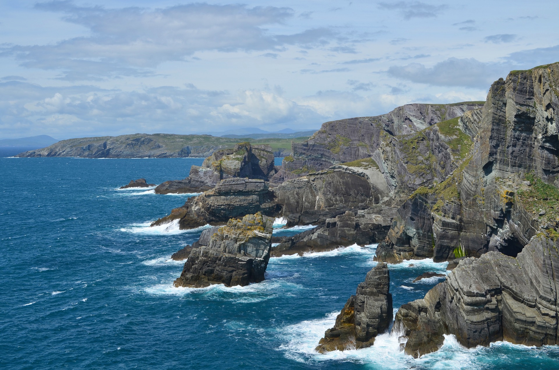 Dramatic rocky cliffs rise above the blue sea with waves crashing at their base under a partly cloudy sky, forming a rugged Cork coastline in Ireland with distant hills in the background.