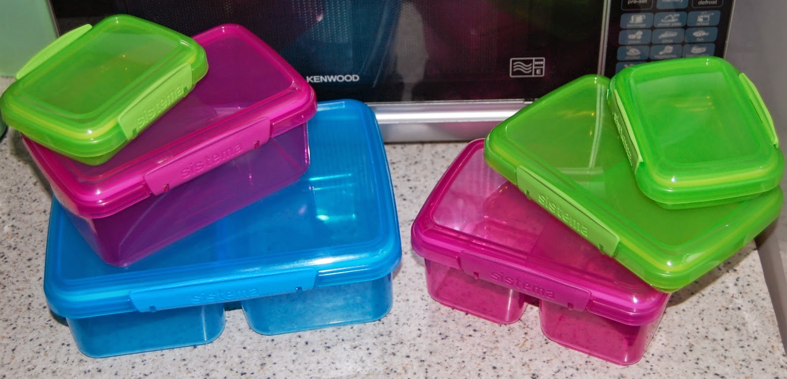 A collection of colourful plastic food containers, perfect for summer leftovers, including blue, pink, and green boxes with matching lids from Ocado, stacked on a kitchen worktop in front of a microwave.
