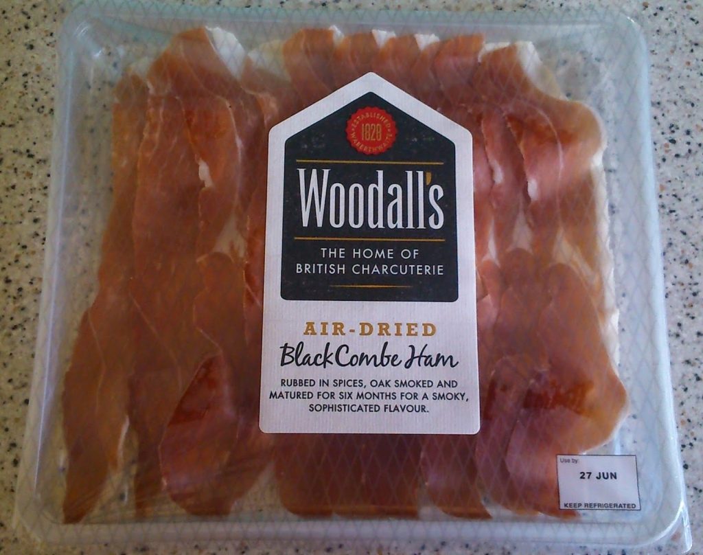 A pack of Woodall's air-dried Black Combe Ham, perfect for summer picnics, with slices visible through the plastic. The label highlights its spice rub, oak smoking, and six-month maturation. Use-by date is marked in the corner. Available at Ocado.