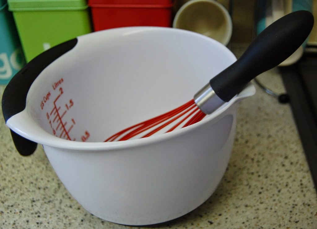 A white mixing bowl with measurement markings and an OXO Good Grips whisk inside, placed on a speckled worktop with colourful containers in the background—essential kitchen tools for every cook.
