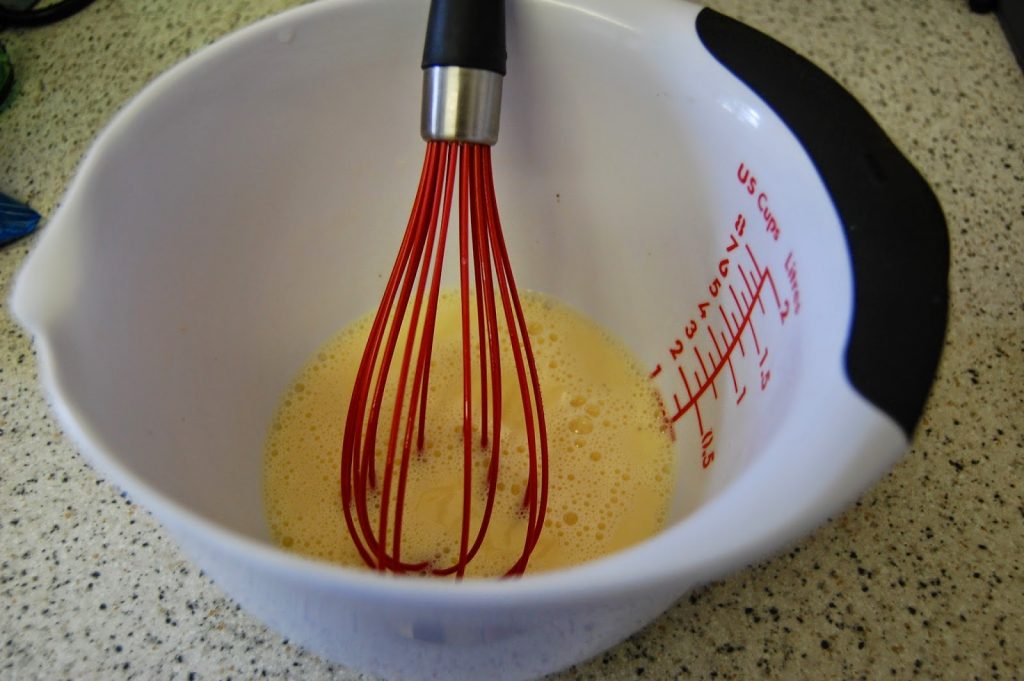 A red OXO Good Grips whisk rests in a white mixing bowl with measurement markings, containing a light yellow, frothy liquid. The bowl sits on a speckled worktop among essential kitchen tools.