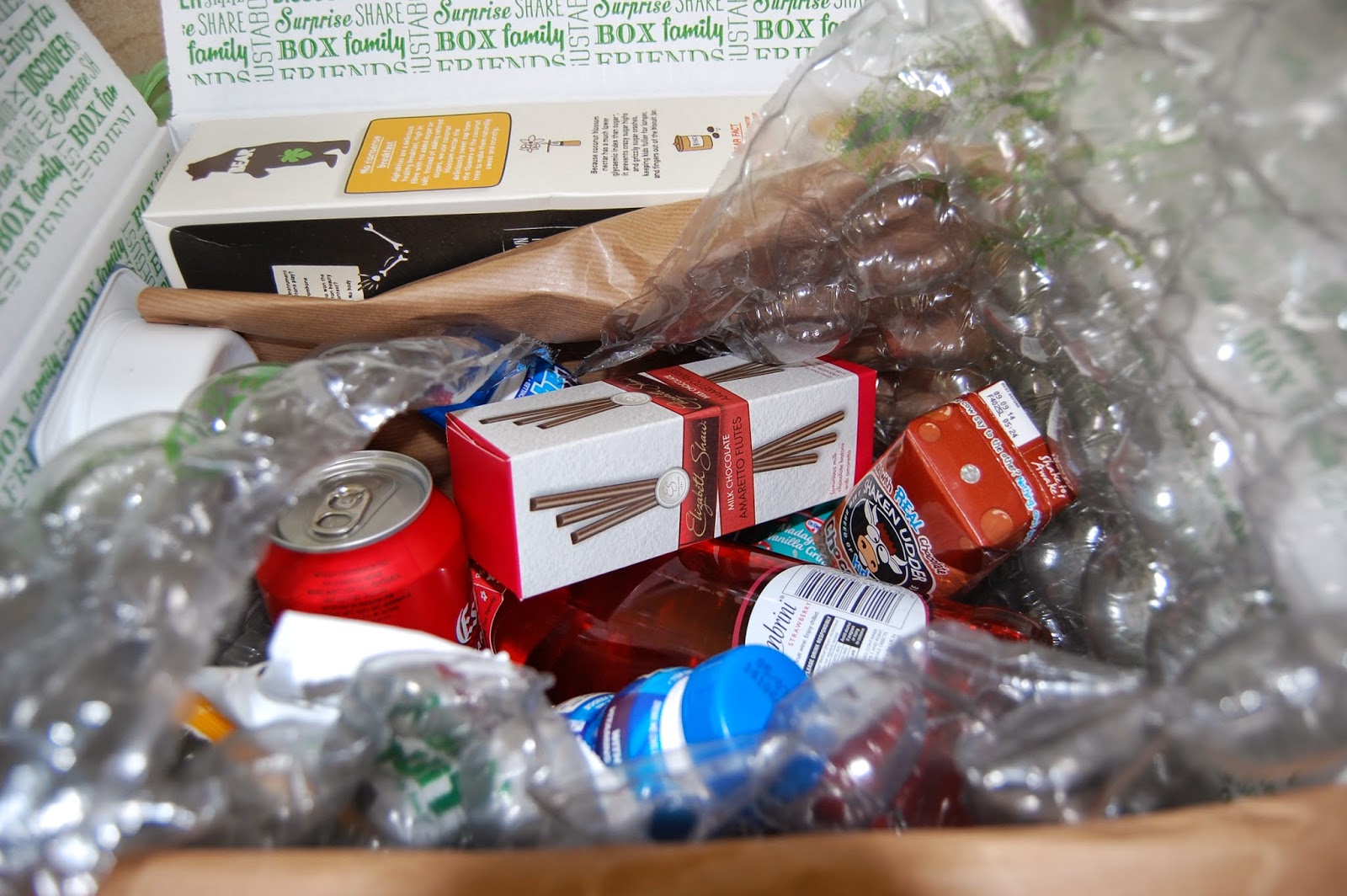 A Degustabox filled with assorted drinks, snacks, and treats, surrounded by bubble wrap for protection. Visible items include fizzy drink cans, boxed biscuits, bottled drinks, and a wooden spoon.